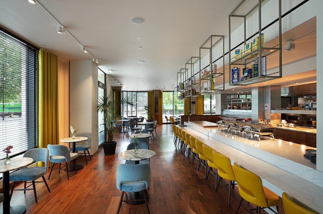 Tower Suites By Blue Orchid London Ngoại thất bức ảnh The photo shows a modern restaurant interior. It features a spacious layout with large windows allowing natural light to flood the space. The decor includes a combination of wooden flooring and colorful furniture—blue and yellow chairs arranged aroun