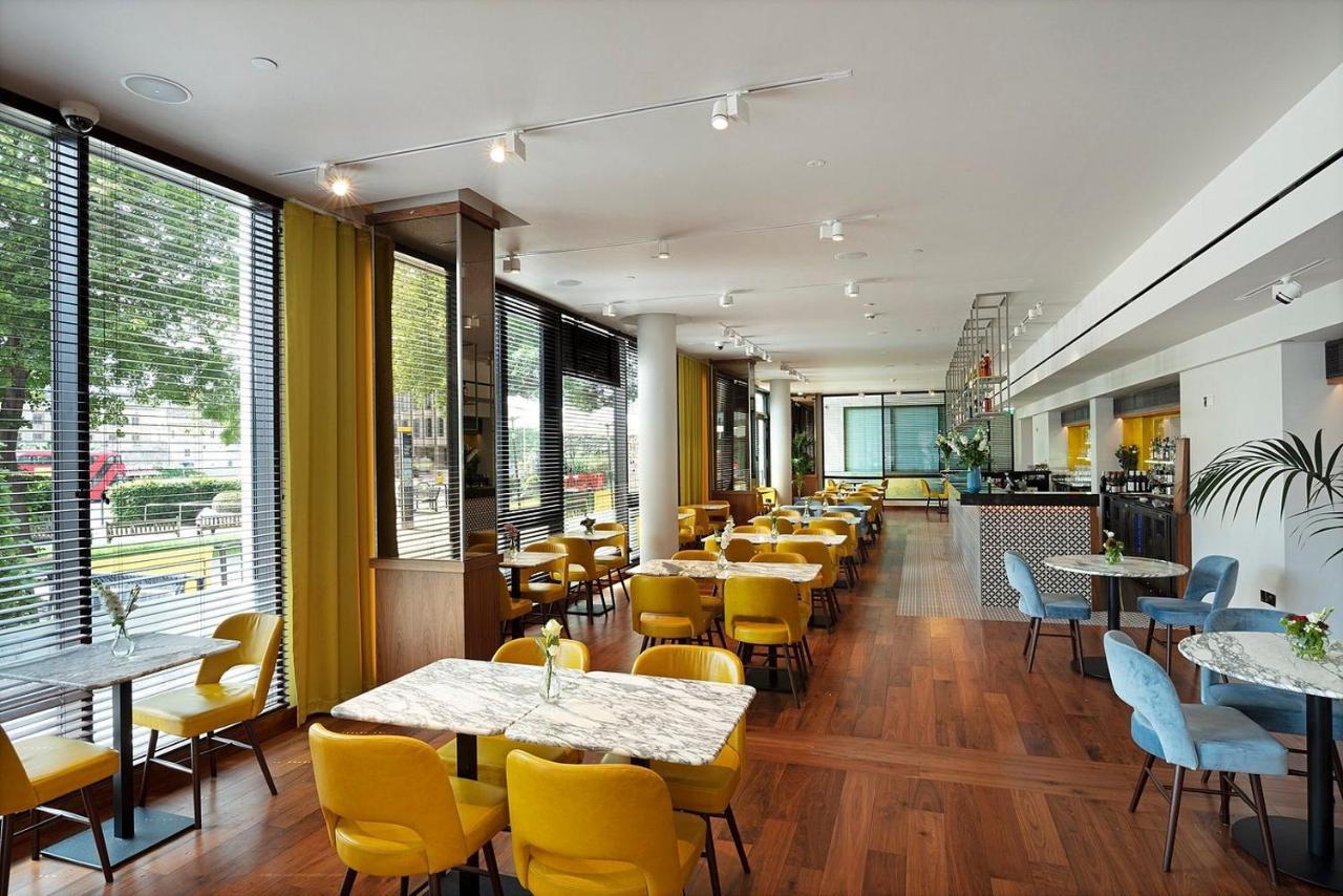 Tower Suites By Blue Orchid London Ngoại thất bức ảnh The photo depicts a modern dining area with a bright and airy atmosphere. There are several tables with marble tops, accompanied by yellow chairs, creating a vibrant contrast with the wood flooring. The space features large windows that allow ample n