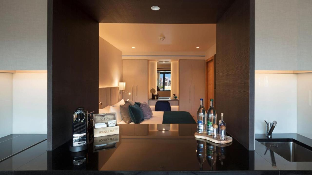 Tower Suites By Blue Orchid London Ngoại thất bức ảnh The image shows a modern hotel room with a stylish design. In the foreground, there is a dark countertop with several bottles displayed, likely including spirits and water. Just beyond the countertop, you can see the bedroom area, which has light-col
