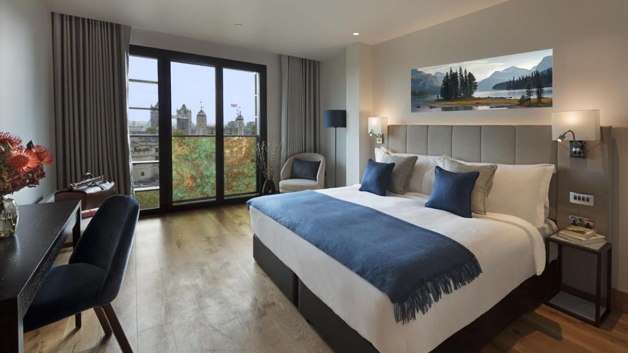 Tower Suites By Blue Orchid London Ngoại thất bức ảnh The photo shows a modern hotel room that features a large bed with white linens and blue accent pillows. There’s a cozy throw blanket at the foot of the bed. The room is well-lit and includes a seating area with a chair and a small table. A large win