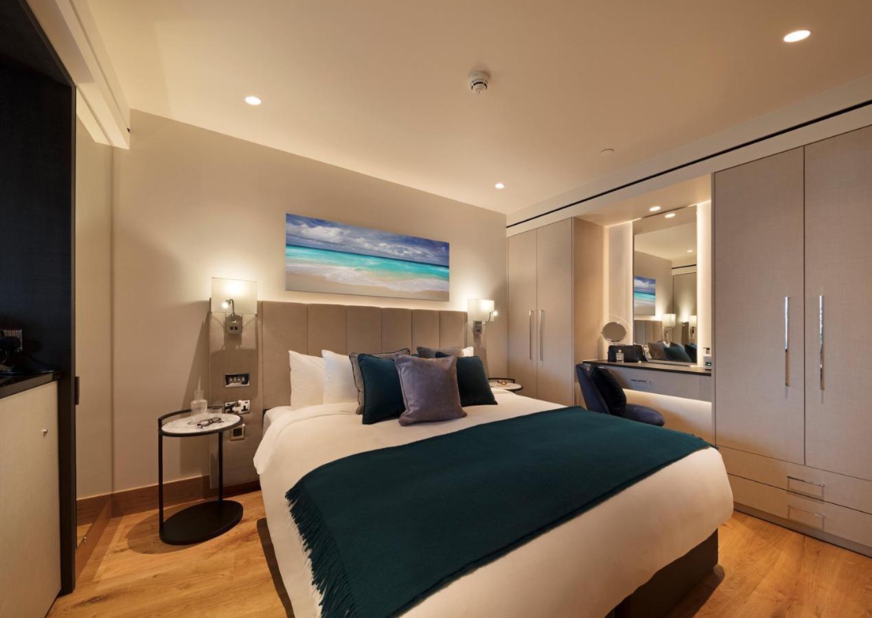 Tower Suites By Blue Orchid London Ngoại thất bức ảnh The photo shows a modern hotel room featuring a double bed with a white duvet and a dark teal throw blanket. There are decorative pillows in shades of blue and purple on the bed. To one side, there's a round bedside table with a small lamp. A large m