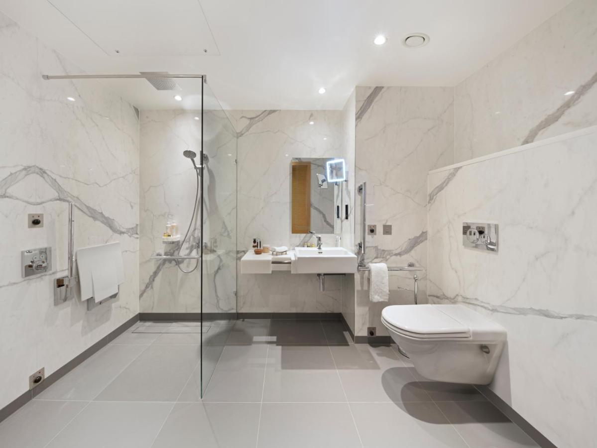 Tower Suites By Blue Orchid London Ngoại thất bức ảnh The photo shows a modern bathroom that features marble walls with a light color. There is a large walk-in shower with a glass partition, and the shower area has a rain shower head. To the right, there is a stylish sink with a mirror above it, and var