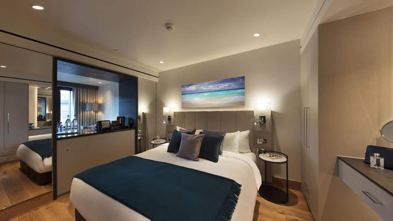 Tower Suites By Blue Orchid London Ngoại thất bức ảnh The photo shows a modern hotel bedroom featuring a large bed with a light-colored headboard and a deep blue throw at the foot. The bed is adorned with multiple decorative pillows. On the wall above the bed, there is a colorful painting depicting a be