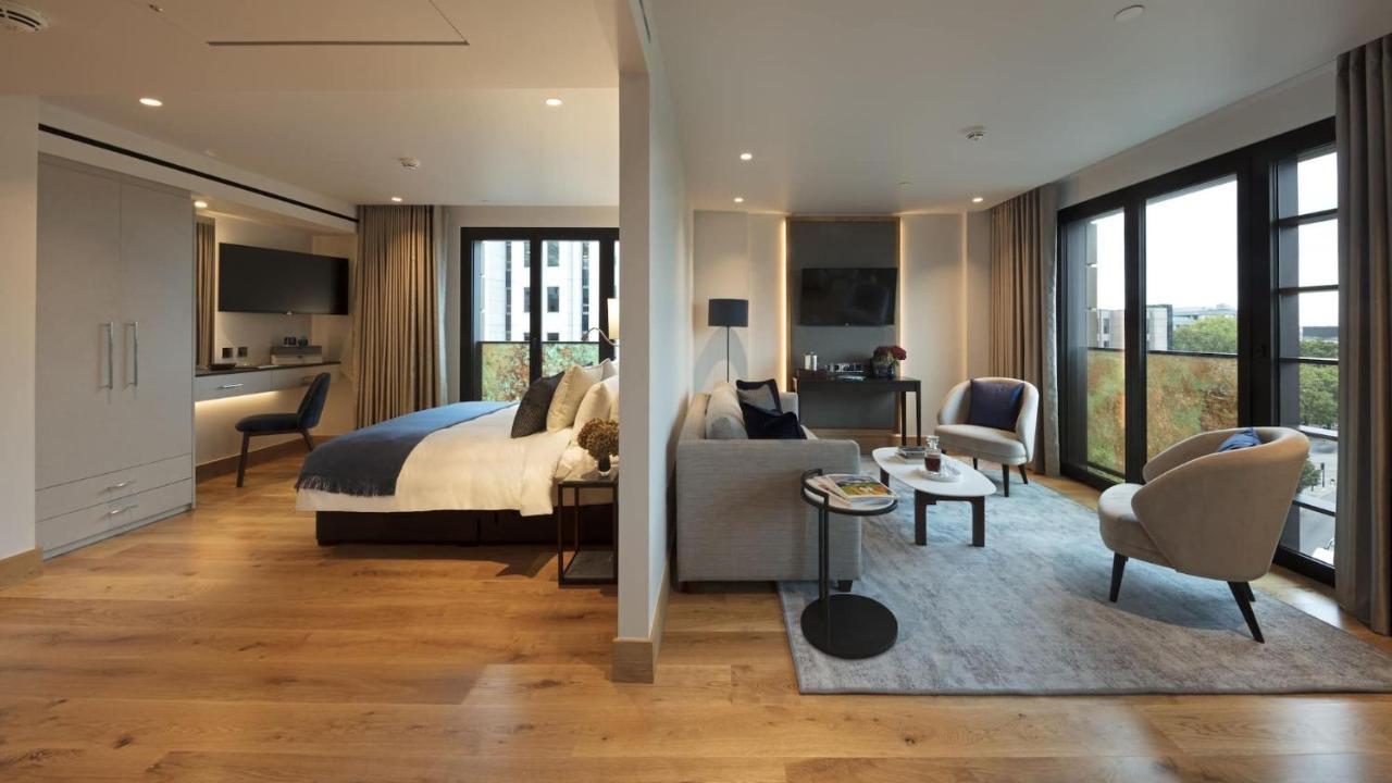 Tower Suites By Blue Orchid London Ngoại thất bức ảnh The photo shows a modern hotel room or suite. On one side, there's a bed with a blue and white duvet and pillows, accompanied by a bedside table with a lamp. Opposite the bed, there's a living area featuring a cozy gray sofa, a small round coffee tab