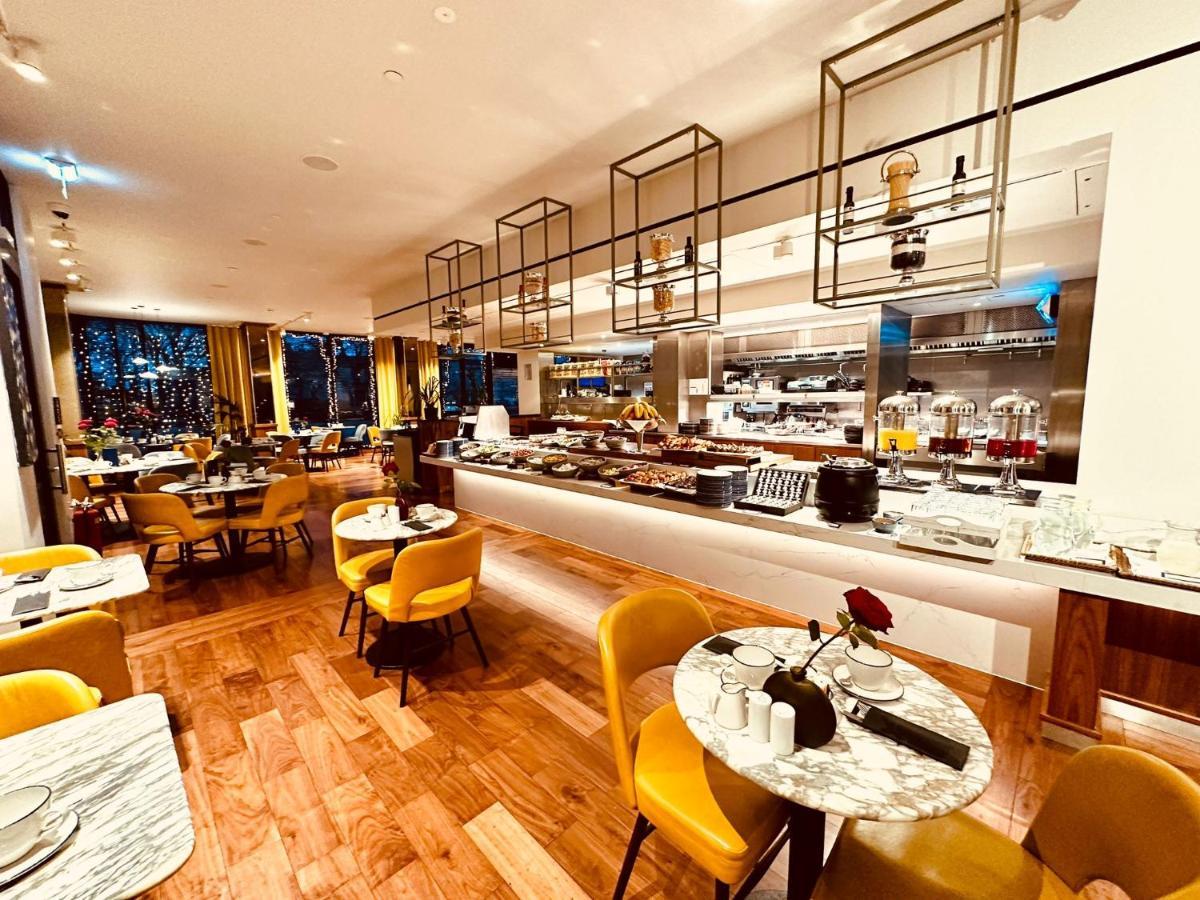 Tower Suites By Blue Orchid London Ngoại thất bức ảnh The photo depicts a modern restaurant interior with a spacious layout. There are several round tables, each adorned with marble tabletops and stylish yellow chairs. A buffet area is visible in the background, featuring various food setups and kitchen