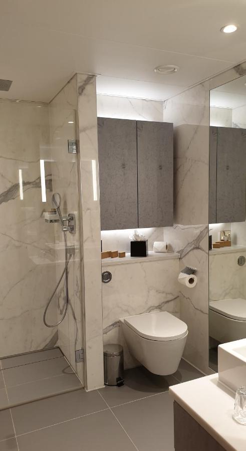 Tower Suites By Blue Orchid London Ngoại thất bức ảnh The photo shows a modern bathroom design featuring elegant marble walls. On one side, there is a transparent shower enclosure with a showerhead and controls visible. Opposite the shower, there is a toilet, and above it are some sleek cabinets. The sp